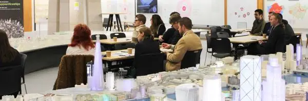 People seated at tables in a exhibition space, listening to a presentation beside a detailed scale model of London with illuminated high-rise buildings and development displays on the walls