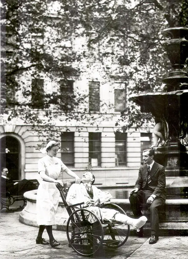 Black and white image of a nurse pushing a wheelchair and man sitting on a fountain in a suit looking at them