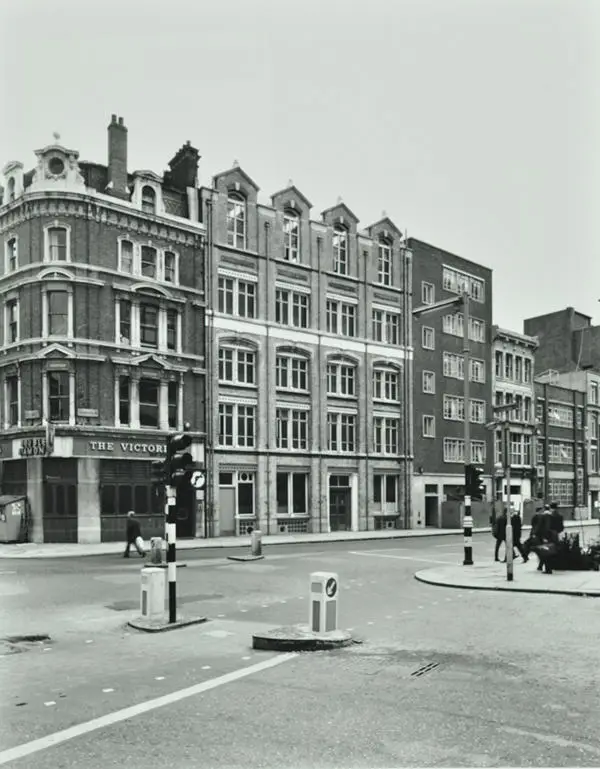 Photograph of street lined with five storey buildings – there are traffic lights on the pavement and several pedestrians
