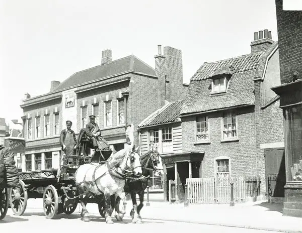 Photograph of a street dominated by a horse-drawn dray wagon with two dray-drivers. Behind them is the two-storey brick pub with a relief of a white horse in the wall’s centre. The stand-alone sign for the Public House includes a statute of a horse. Besides the pub are two smaller two-storey houses