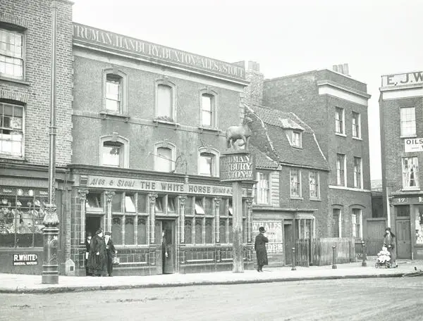 Photograph of a pub surrounded by other three-storey buildings. The ground floor is decorated in Victorian glazing. There is a stand alone sign topped by statuette of a horse. Three individuals are exiting the Public House, with another smoking in the doorway. Two pedestrians, one with a baby in a pushchair walk in opposite directions
